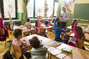 Happy elementary teacher asked the question while group of children raised their hands ready to answer it.